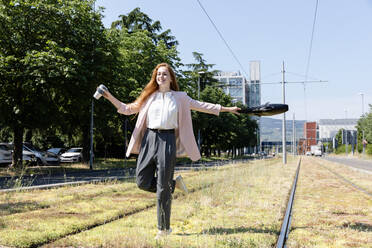 Lächelnde weibliche Fachkraft, die wegschaut, während sie eine Tasche inmitten von Eisenbahnschienen schwingt - EIF01529