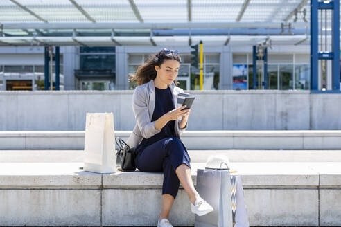 Frau mit Einkaufstüten, die ihr Smartphone benutzt, während sie auf einer Stützmauer sitzt - EIF01583