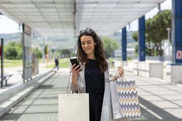Woman using smart phone carrying shopping bags at tram station - EIF01589