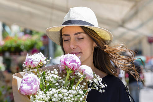 Frau mit Sonnenhut riecht an Pfingstrosen Blumenstrauß in der Nähe von Blumenladen - EIF01611