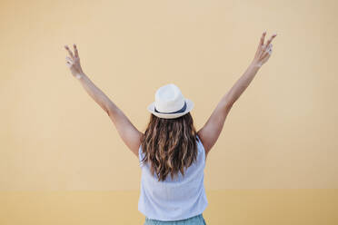 Woman in hat gesturing peace sign while standing in front of brown wall - EBBF04177