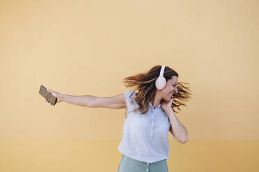Cheerful woman with mobile phone listening music through headphones against wall - EBBF04183
