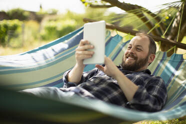 Glücklicher Mann mit digitalem Tablet in sonniger Sommerhängematte - CAIF30868