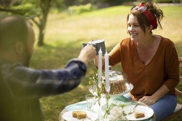 Glückliches Paar genießt Tee und Kuchen am Tisch im sonnigen Garten - CAIF30886