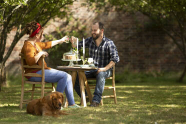 Happy couple with dog toasting glasses at table in summer garden - CAIF30904