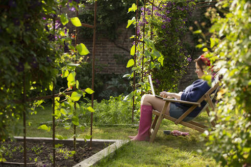 Frau benutzt Laptop im sonnigen Sommergarten - CAIF30916