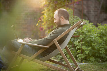 Man working at laptop in lawn chair in summer garden - CAIF30923