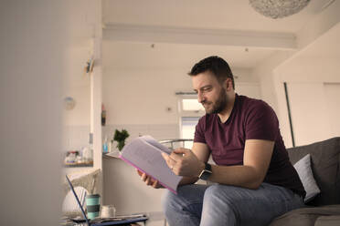 Man with paperwork working from home at laptop on sofa - CAIF30987