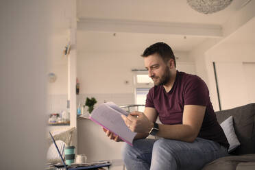 Mann mit Papierkram, der von zu Hause aus am Laptop auf dem Sofa arbeitet - CAIF30987