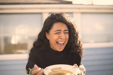 Fröhlich lachende Frau, die auf einer sonnigen Veranda Eintopf isst - CAIF30988