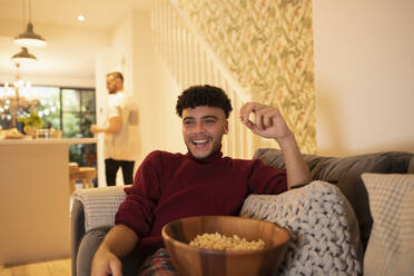 Glücklicher junger Mann mit Popcorn vor dem Fernseher auf dem Wohnzimmersofa - CAIF30994