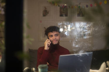 Junger Mann arbeitet von zu Hause aus und spricht mit seinem Smartphone am Laptop - CAIF31014