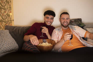 Glückliches schwules männliches Paar mit Wein und Popcorn, das auf dem Sofa einen Film schaut - CAIF31039