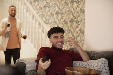 Happy young man with remote control watching TV on sofa - CAIF31044
