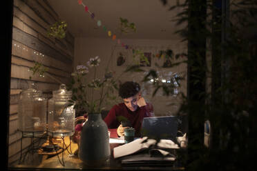 Junger Mann mit Smartphone arbeitet nachts von zu Hause aus am Laptop - CAIF31048