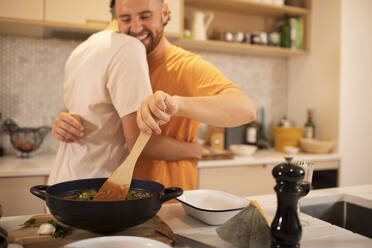 Happy gay male couple hugging and cooking in kitchen - CAIF31049