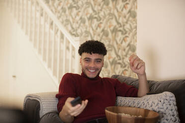 Happy young man with remote control watching TV on sofa - CAIF31050