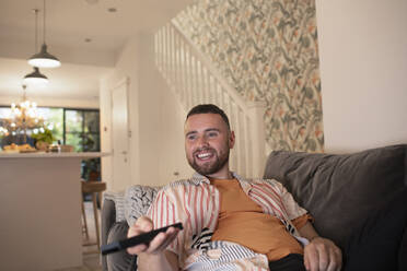 Happy young man with remote control watching TV on sofa - CAIF31065