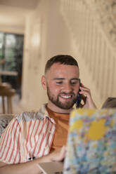 Smiling young man talking on smart phone at laptop - CAIF31076