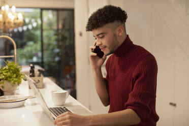 Junger Mann arbeitet von zu Hause aus und spricht mit seinem Smartphone am Laptop - CAIF31084