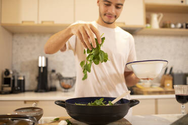 Junger Mann beim Kochen mit frischem Spinat in der Küche - CAIF31120