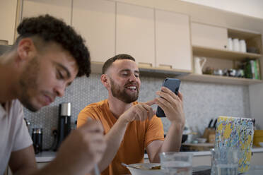 Glückliches schwules männliches Paar, das ein Smartphone benutzt und in der Küche isst - CAIF31127