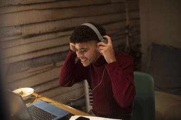 Happy young man with headphones working from home at laptop - CAIF31130