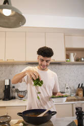 Lächelnder junger Mann beim Kochen mit frischem Basilikum in der Küche - CAIF31134