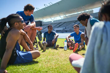 Junge männliche Sportlerfreunde, die sich auf dem sonnigen Rasen des Stadions ausruhen und unterhalten - CAIF31196