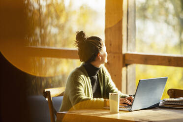 Woman working at laptop looking out window in sunny cafe - CAIF31220