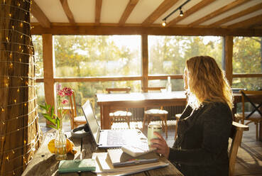 Nachdenkliche Geschäftsfrau arbeitet am Laptop in einem sonnigen Restaurant - CAIF31234