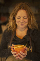 Glückliche weibliche Barista hält Cappuccino mit herzförmigem Schaum - CAIF31275