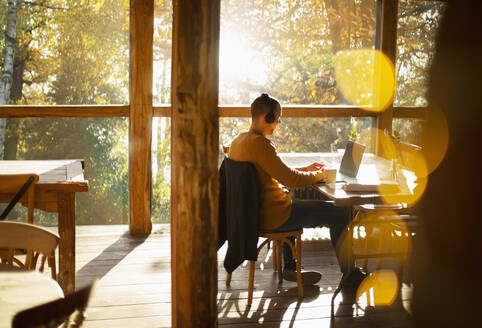 Geschäftsmann mit Kopfhörern arbeitet am Laptop in einem sonnigen Café - CAIF31278