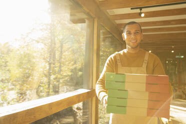 Porträt stolzer männlicher Pizzeriabesitzer mit Pizzakartons am sonnigen Fenster - CAIF31283