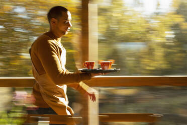 Männlicher Barista trägt Tablett mit Kaffeetassen am Fenster eines Herbstcafés entlang - CAIF31289