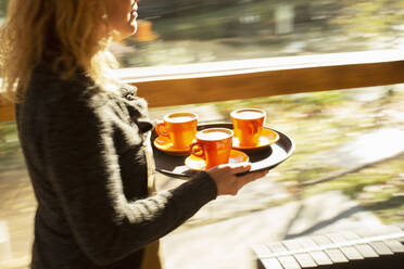 Eine Barista trägt ein Tablett mit Kaffeetassen am Fenster eines sonnigen Cafés entlang - CAIF31290