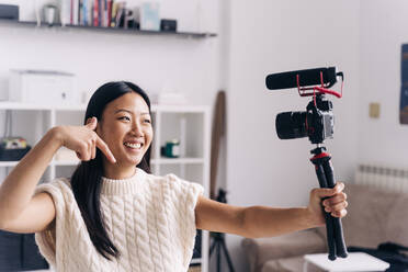 Smiling ethnic female vlogger recording video on photo camera while making hand gestures standing in living room - ADSF25408