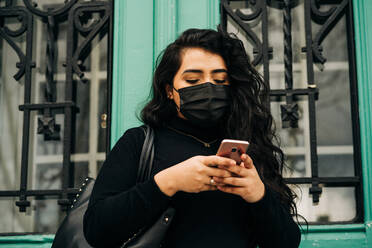 Niedriger Blickwinkel einer seriösen Frau mit Schutzmaske, die im Internet auf ihrem Smartphone surft, während sie auf der Straße in der Nähe eines Gebäudes steht, das vom Coronavirus befallen ist - ADSF25416