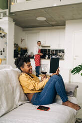 Fröhliche afroamerikanische Frau liest auf dem Sofa ein interessantes Buch vor dem Hintergrund eines Mannes, der in der Küche kocht - ADSF25425