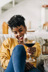 Zufriedene afroamerikanische Frau sitzt auf dem Sofa mit einem Glas Rotwein und genießt das Wochenende zu Hause, während sie in die Kamera schaut - ADSF25427
