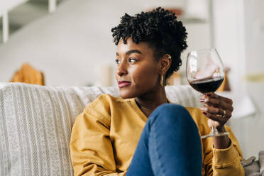 Content African American female sitting on sofa with glass of red wine and enjoying weekend at home while looking away - ADSF25428