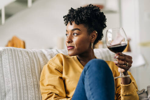 Zufriedene afroamerikanische Frau sitzt auf dem Sofa mit einem Glas Rotwein und genießt das Wochenende zu Hause, während sie wegschaut - ADSF25428
