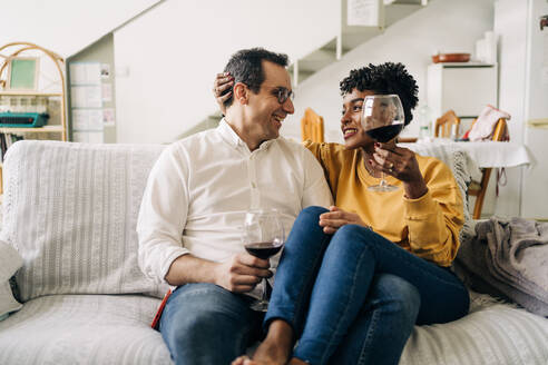 Zufriedenes multirassisches Paar, das auf dem Sofa zu Hause mit Rotwein in Gläsern chillt, während es das Wochenende zu Hause genießt und sich gegenseitig anschaut - ADSF25429