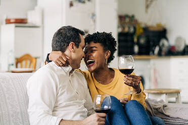 Zufriedenes multirassisches Paar, das auf dem Sofa zu Hause mit Rotwein in Gläsern chillt, während es das Wochenende zu Hause genießt und sich gegenseitig anschaut - ADSF25430