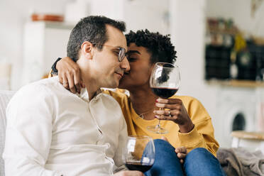 Zufriedenes multirassisches Paar, das auf dem Sofa zu Hause mit Rotwein in Gläsern chillt, während es das Wochenende zu Hause genießt und sich küsst - ADSF25431