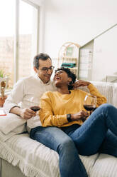 Content multiracial couple chilling on sofa at home with red wine in glasses while enjoying weekend at home and looking at each other - ADSF25432