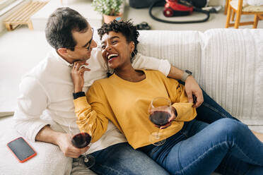Zufriedenes multirassisches Paar, das auf dem Sofa zu Hause mit Rotwein in Gläsern chillt, während es das Wochenende zu Hause genießt und sich gegenseitig anschaut - ADSF25433