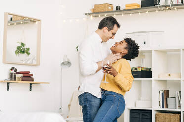 Side view of smiling multiethnic couple in love standing with glass of wine and embraced while spending romantic weekend at home and looking at each other - ADSF25437