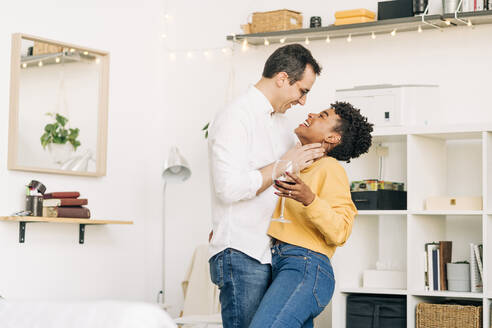 Seitenansicht eines lächelnden multiethnischen verliebten Paares, das mit einem Glas Wein dasteht und sich umarmt, während es ein romantisches Wochenende zu Hause verbringt und sich gegenseitig anschaut - ADSF25437