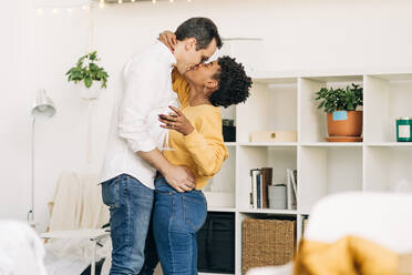 Seitenansicht eines lächelnden multiethnischen verliebten Paares, das mit einem Glas Wein steht und sich küsst, während es ein romantisches Wochenende zu Hause verbringt - ADSF25439